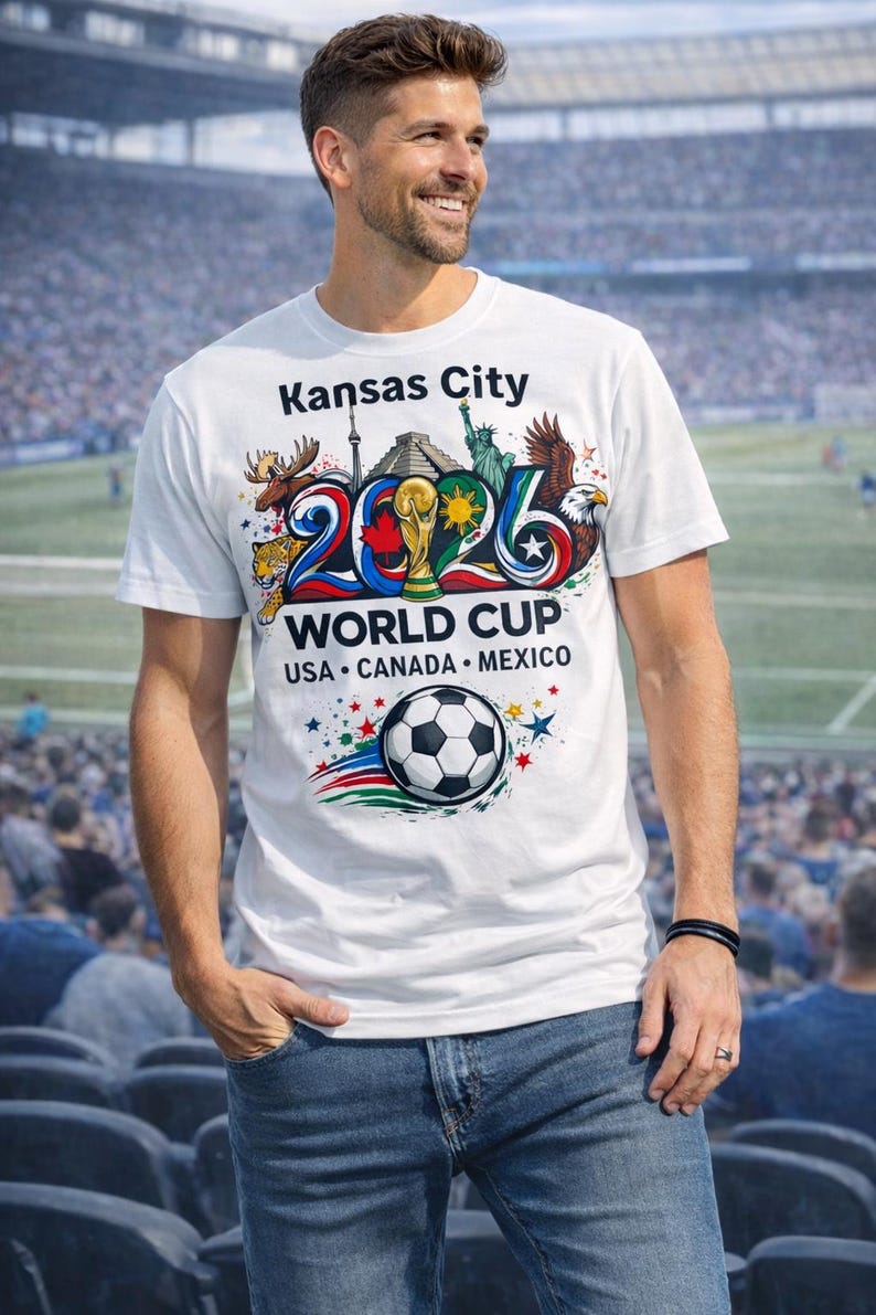 May include: White t-shirt with a colorful graphic promoting the 2026 World Cup in Kansas City, USA, Canada, and Mexico. The design includes a soccer ball, the World Cup trophy, and various national symbols.