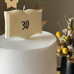 Peut inclure: Une bougie blanche en forme de livre avec le chiffre 30 imprimé en noir sur la couverture. La bougie est placée verticalement sur un gâteau cylindrique blanc lisse, avec une mèche blanche unique dépassant du centre supérieur du livre.