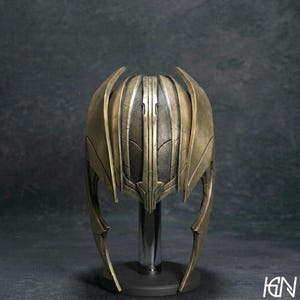 May include: A metallic gold and dark bronze helmet featuring a multi-paneled design with vertical ridges and curved side extensions. The helmet has a weathered, textured finish and is mounted on a central vertical stand for display purposes.