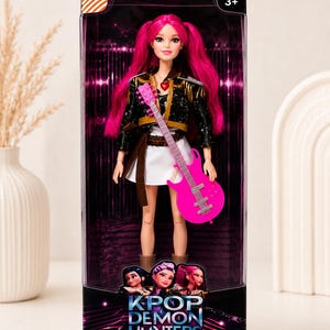 May include: A doll with bright pink hair and a matching guitar, dressed in a black jacket and white skirt. The doll is packaged in a box with the text "KPOP DEMON HUNTERS" and the tagline "COLLECT. SING. SLAY."