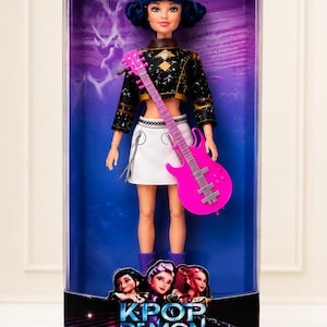 May include: A doll in a clear plastic box. The doll has blue hair, a black and gold top, a white skirt, and purple boots. She holds a pink guitar. The box reads "KPOP DEMON HUNTERS" and "COLLECT. SING. SLAY."