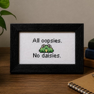 May include: A black rectangular frame containing a white cross-stitch fabric piece. The embroidery features a green pixelated frog centered between the black text "All oopsies." above and "No daisies." below, creating a humorous design.