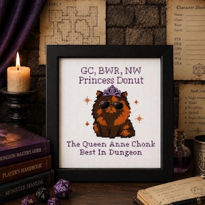 May include: A framed cross-stitch featuring a cartoon cat wearing sunglasses and a crown. The text reads "GC, BWR, NW Princess Donut" and "The Queen Anne Chonk Best In Dungeon." The image is surrounded by books, dice, and a lit candle.