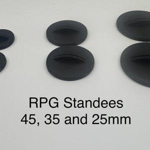 May include: Six black circular RPG standees with a central slot for game pieces, arranged in three pairs of varying sizes. The text below reads: RPG Standees 45, 35 and 25mm. These round bases are designed to hold tabletop gaming miniatures securely.