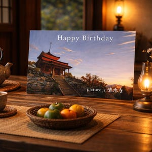 May include: A birthday card with a Japanese temple scene, the text "Happy Birthday" is at the top. The card is on a wooden table with a teapot, cup, and a basket of fruit. A lit lamp and vase are also on the table.