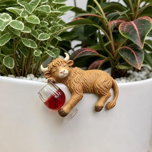 Puede incluir: Una figura caprichosa de una vaca de las Tierras Altas descansando junto a una copa de vino en miniatura llena de líquido rojo. La vaca es de color marrón claro con pequeños cuernos y una cara sonriente. La copa es transparente y la escena se desarrolla sobre plantas verdes.