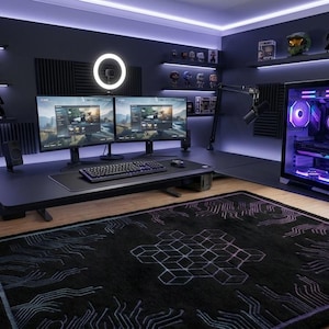Op de afbeelding: Een zwart rechthoekig vloerkleed met een wit geometrisch printpatroon van printplaten met paarse en blauwe accenten. Het kleed ligt in een kamer met een gamingbureau, twee monitoren, een computerkast met paarse verlichting en wandplanken.