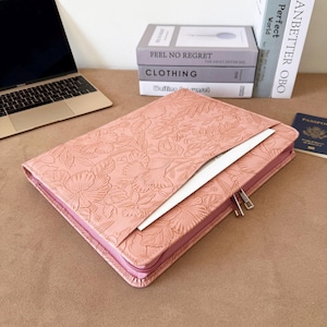 May include: A rectangular, dusty pink laptop sleeve featuring an embossed floral pattern across the exterior. The sleeve has a zippered closure and a front pocket, with a silver-colored zipper pull visible on the side of the case.