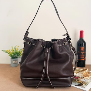 Pode incluir: Uma bolsa saco castanho-escura com fecho de cordão com dois puxadores de couro compridos, ferragens de metal em tom de latão e costuras brancas contrastantes ao longo das costuras e da base. A bolsa inclui uma alça de ombro única.