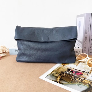 May include: A dark gray leather clutch bag with a pebbled texture and a fold-over flap closure. The rectangular bag features a simple, clean design with a centered circular detail on the flap, providing a structured yet soft appearance for storage.