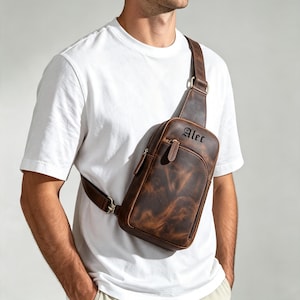 May include: A brown leather crossbody sling bag featuring a single adjustable shoulder strap and a front zippered pocket. The bag is embossed with the name Alec in a black gothic-style font on the upper front section of the leather surface.