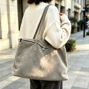 Op de afbeelding: Een grote, lichtgrijze suède tote bag met twee schouderbanden en een centrale draaisluiting van zilverkleurig metaal. De tas heeft een zachte, matte textuur en een gestructureerde, rechthoekige vorm, gedragen over de schouder.