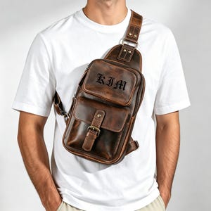 Puede incluir: Un bolso bandolera de cuero marrón que presenta un compartimento superior con cremallera con el nombre KIM grabado en fuente gótica, un bolsillo inferior con cierre de hebilla y una correa ajustable ancha con detalles metálicos.