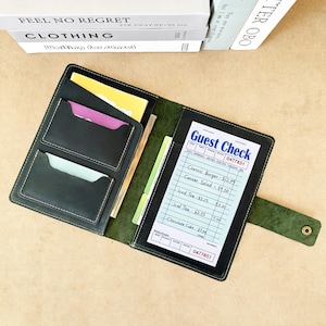 May include: A dark green leather server book featuring a snap closure, multiple interior pockets for cards, and a clear sleeve holding a white Guest Check pad with printed text including Classic Burger, Caesar Salad, Iced Tea, and Chocolate Cake.