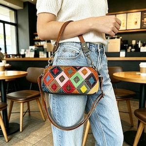 May include: A cylindrical handbag featuring a quilted diamond pattern in various colors including red, blue, green, pink, and yellow. The bag has a brown leather handle and a detachable shoulder strap with metal hardware, worn against light blue jeans.
