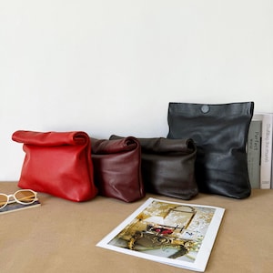 May include: Four leather pouches in red, burgundy, dark brown, and black, each featuring a roll-top design with a textured surface. The pouches are arranged in a row, with the black pouch positioned furthest to the right.