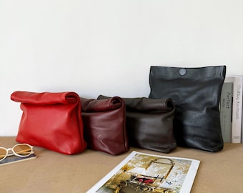 May include: Four roll-top clutch bags in red, burgundy, dark brown, and black leather with a pebbled texture. Each bag features a simple fold-over design. A pair of white-rimmed sunglasses and a printed paper sheet are placed in the foreground.