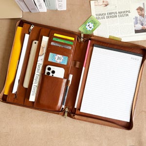 May include: Open brown leather portfolio with various compartments, including card slots, a phone pocket, and a notepad. The portfolio contains a pen, a yellow document, and a white notepad with the word "MEMO" printed on it.