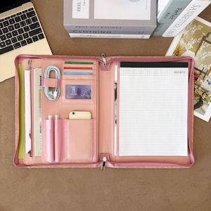 May include: A pink zippered portfolio organizer featuring multiple interior slots for cards, a clear ID window, two pen loops, a smartphone pocket, a coiled light blue charging cable, and a lined memo pad with a grid header on the right side.