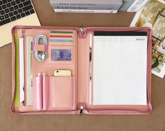 May include: A pink zippered portfolio organizer featuring multiple interior slots for cards, a clear ID window, two pen loops, a smartphone pocket, a coiled light blue charging cable, and a lined memo pad with a grid header on the right side.
