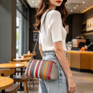 May include: A crossbody bag featuring vertical stripes in shades of red, orange, yellow, green, and blue, with a solid gray side panel and a matching gray shoulder strap, worn with a white ribbed short-sleeve top and light blue denim jeans.