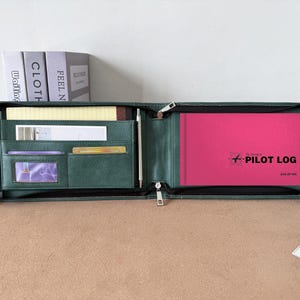 Könnte beinhalten: Eine dunkelgrüne Reißverschluss-Organizer-Mappe mit mehreren Innentaschen für Karten und Dokumente, einer Stiftschlaufe und einem leuchtend pinkfarbenen Flugbuch mit dem Aufdruck "The Standard PILOT LOG" und "ASA-SP-6A" auf dem Einband.