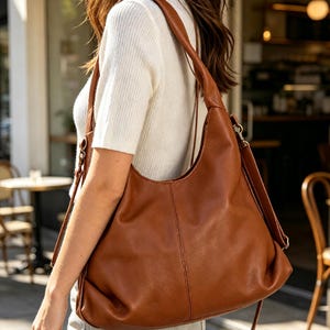 May include: A large, slouchy hobo-style shoulder bag in a rich cognac brown color. The bag features a smooth, matte finish, a single wide shoulder strap, and a vertical seam detail running down the center of the front panel with metallic hardware.