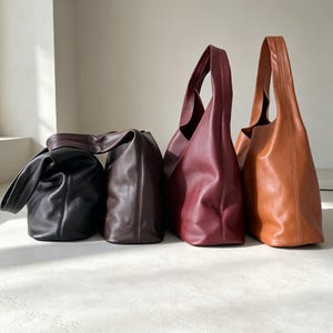 May include: Four hobo bags arranged in a row, featuring a smooth finish and a single shoulder strap. The bags are colored in black, dark brown, deep burgundy, and tan. Each bag has a slouchy, unstructured shape with visible vertical seams.