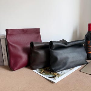 May include: Three leather bags in varying sizes and colors. A large, flat, burgundy bag stands upright. Two smaller bags, one brown and one black, have a rolled-over top. The bags are on a neutral surface with a bottle of wine in the background.