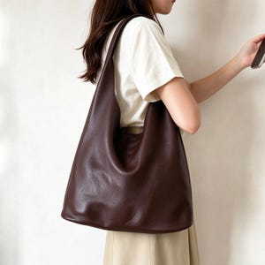 May include: A large, dark brown hobo-style shoulder bag with a smooth, matte texture. The bag features a wide, integrated shoulder strap and a spacious, slouchy main compartment that hangs vertically against the side of the body.