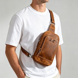 Puede incluir: Un bolso bandolera de cuero color canela que se lleva cruzado sobre el pecho, con un bolsillo frontal con cremallera y el monograma A.E. grabado en negro. El bolso tiene una correa ajustable con herrajes de metal plateado y acabado mate.