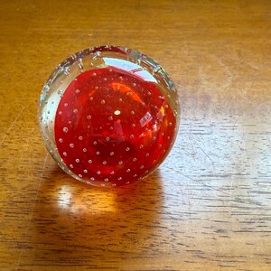 May include: A clear glass paperweight with a vibrant red core. The red core is speckled with small, clear bubbles, and the outer glass is also dotted with bubbles. The paperweight is spherical and sits on a wooden surface.