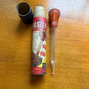 Könnte beinhalten: Ein Artbeck Baster-Set mit einem roten und klaren Glasbaster, einer Pappröhre und einem zylindrischen Behälter. Der Behälter hat Text und Illustrationen, darunter "Artbeck Baster" und Gebrauchsanweisungen. Der Baster hat eine rote Glühbirne und ein klares Glasrohr.
