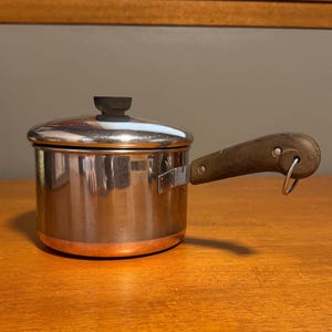May include: A stainless steel saucepan with a copper-colored base, a matching lid with a dark knob, and a brown handle featuring a metal hanging loop. The pot has a cylindrical body and a side-mounted handle attached with visible metal rivets.