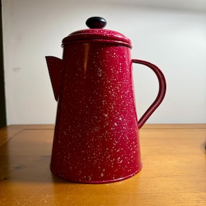 May include: A red enamel coffee pot with white speckles. The pot has a black knob on the lid and a curved handle. The spout is on the left side. The pot is on a wooden surface.