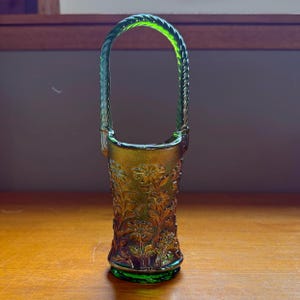 May include: A green glass basket with a braided handle. The basket is decorated with a floral pattern and has a round base. The glass has an iridescent finish, giving it a shimmering appearance. The basket is empty and sits on a wooden surface.