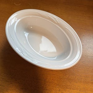 May include: A white oval ceramic bowl featuring a smooth, glossy finish and a subtle, embossed leaf pattern along the wide, rounded rim. The bowl has a deep, concave center and a clean, minimalist design with no additional colors or markings.