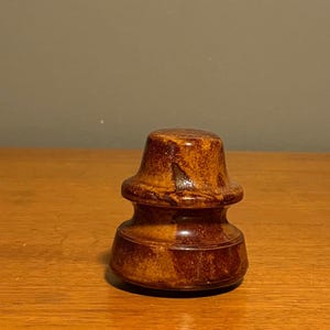 Può includere: Un isolatore elettrico in ceramica marrone con una finitura lucida e maculata. L'oggetto presenta un design a gradini a forma di campana con una parte superiore piatta e una base circolare più ampia, con sfumature di marrone chiaro e scuro.
