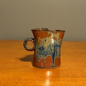 Puede incluir: Una jarrita de cerámica para crema con un esmalte base de color terracota cálido y un patrón de goteo vertical prominente en tonos azul, crema y beige. El recipiente tiene un asa circular pequeña y un pico vertedor distintivo en el borde.