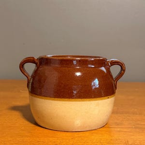 May include: A two-handled ceramic pot featuring a glossy dark brown glaze on the upper portion and a matte cream-colored base. The vessel has a wide, rounded body with two curved handles extending from the sides near the top rim.