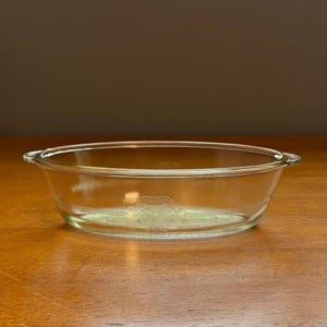 May include: A clear glass oval baking dish featuring integrated side handles and a decorative etched pattern around the exterior rim. The design consists of symmetrical scrollwork and horizontal lines embossed into the transparent glass surface.