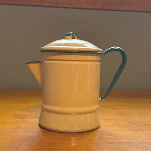 以下が含まれることがあります： クリーム色のエナメル製コーヒーポットで、縁、ハンドル、蓋のつまみはダークグリーンです。円筒形の容器は幅広の底、短い注ぎ口、小さな取っ手付きの蓋が特徴です。外側全体が滑らかで光沢のある仕上がりになっています。