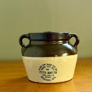 May include: A two-handled stoneware crock featuring a dark brown glazed upper section and a cream-colored base. The front displays blue text reading: WHEN YOU BAKE TRY ME WHEN YOU BUY TRY PETER BOOTZIN THE CORFER STORE MEDFORD, WIS.
