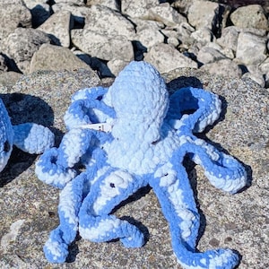 Puede incluir: Un pulpo de peluche hecho a mano, en tonos azul claro y blanco. El pulpo tiene ocho largos tentáculos rizados y una cabeza redondeada. El material suave y texturizado le da un aspecto tierno. El pulpo está sobre una roca gris.