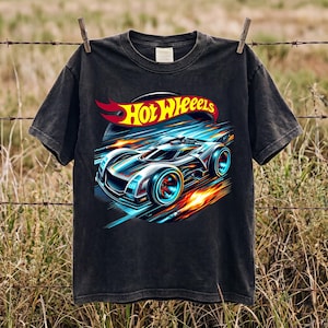 Può includere: Una t-shirt nera a maniche corte con una stampa grafica di un'auto da corsa stilizzata in movimento con scie luminose blu e arancioni. Il logo Hot Wheels è stampato in giallo e rosso sopra l'auto. La maglietta ha un girocollo.
