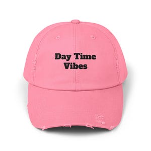 May include: A pink baseball cap with a distressed brim. The text "Day Time Vibes" is printed in black across the front of the cap. The cap is made of a soft material and has a curved brim.