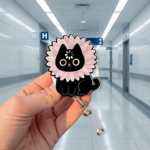 Puede incluir: Un portatarjetas con un gato negro y un diseño floral rosa alrededor de la cabeza. El gato tiene ojos grandes y redondos y una nariz pequeña. El portatarjetas tiene una correa de plástico transparente y un clip metálico.