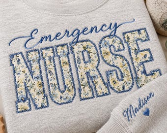 May include: A light gray crewneck sweatshirt featuring the embroidered text Emergency NURSE in blue cursive and block letters. The block letters are filled with a floral pattern of blue and yellow flowers. The name Madison is embroidered on the sleeve.