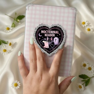 May include: A clear protective tablet case featuring a heart-shaped sticker with a black barbed wire border. The sticker displays a bat holding a scroll, a tombstone, a crescent moon, and the text NOCTURNAL READER in a light pink colour.