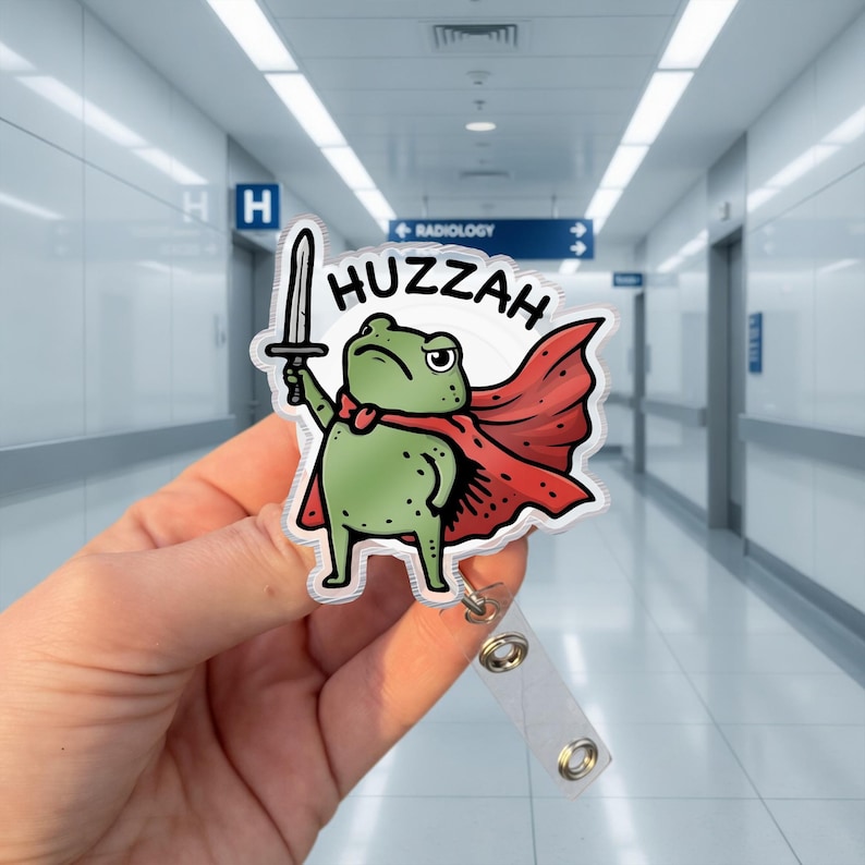 May include: A badge reel featuring a cartoon frog with a sword, red cape, and the word "Huzzah". The frog is green with black spots and a grumpy expression. The reel has a clear plastic strap and a metal clip.